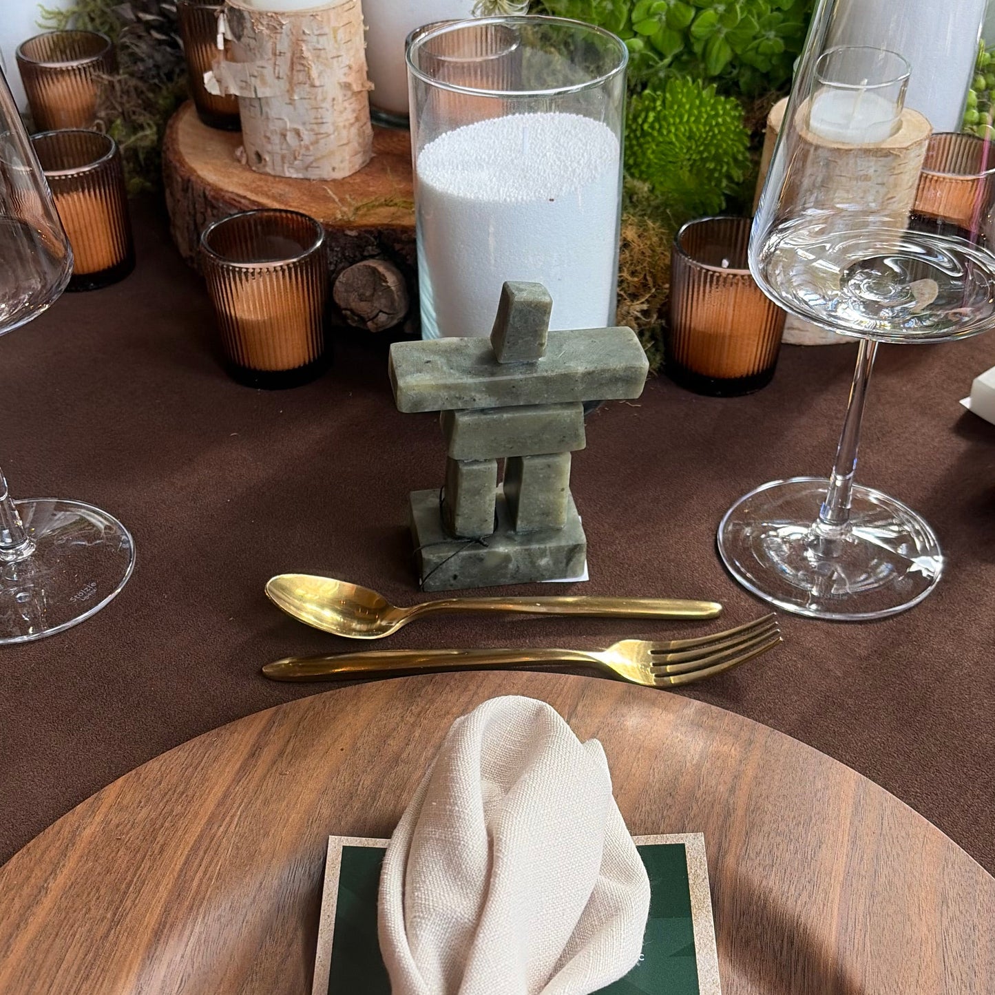 A stone inukshuk on a styled dining table setting featuring a large round wooden charger plate with a cream napkin and a brown leather ring, alongside clear glass candle holders, gold flatware, and green foliage on a dark brown tablecloth.