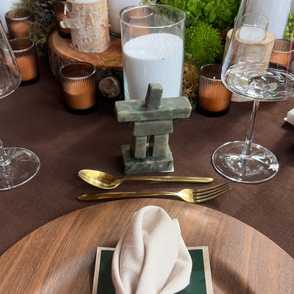 A stone inukshuk on a styled dining table setting featuring a large round wooden charger plate with a cream napkin and a brown leather ring, alongside clear glass candle holders, gold flatware, and green foliage on a dark brown tablecloth.