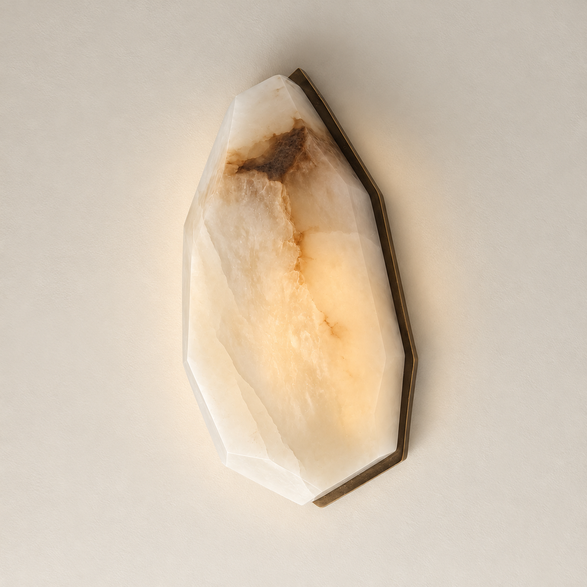 A faceted crystal-style alabaster wall sconce made from natural stone, glowing with warm light from within. The stone has a cream and off-white base with warm tan and brown mineral variation, mounted on a dark metal base against a plain light beige wall.