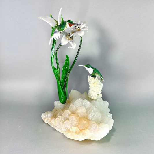 This exquisite sculpture, titled "Hummingbirds," captures the delicate beauty of nature through masterful stone craftsmanship. The piece features two vibrant hummingbirds poised around a pair of blooming lilies, each element meticulously carved from a variety of stones. The birds are brought to life with bodies of agate and jade, accented by howlite and British Columbia red jasper, with eyes of smoky quartz and feet of black jade. The lilies' petals are made of jadeite, while the stems and leaves are skillf