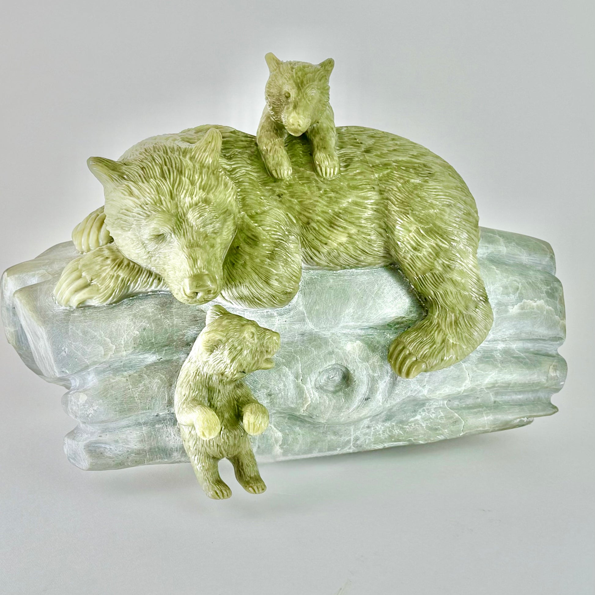Old Growth" is a heartwarming sculpture carved from Australian green chlorite, depicting a serene mother bear with her playful cubs. The artist has skillfully utilized the stone’s natural variations to highlight the tender interaction among the bears. Measuring 15" x 10" x 11" and weighing 55 pounds, this piece exudes a sense of tranquility and familial warmth. The polished green chlorite brings a lifelike vibrancy to the bears, while the log they rest on is rendered with a softer, more muted finish, creati
