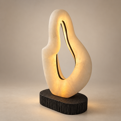 A modern abstract sculptural stone table lamp with a warm glowing cream-colored organic looped body mounted on a dark textured oval base, set against a neutral beige background.
