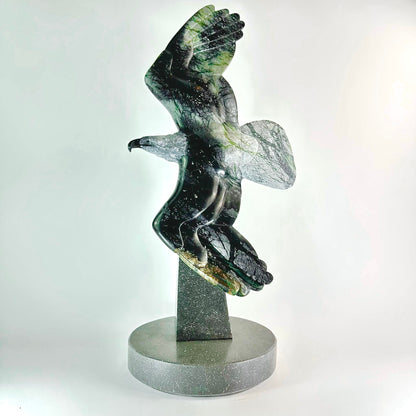 This remarkable stone carving showcases the elegance of a majestic eagle in flight, rendered in authentic BC Green &amp; Black Brucite. The mineral's contrasting palette—deep charcoal accented with vibrant jade striations—creates a visually compelling piece that captures movement and grace. Hand-sculpted to emphasize the stone's natural patterns, each artwork stands as a singular creation. Ideal for collectors, nature enthusiasts, and those who appreciate fine craftsmanship, this sculpture transforms any en