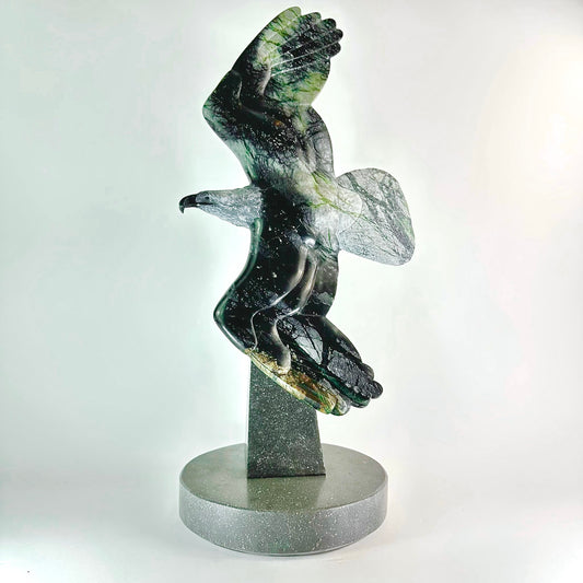 This remarkable stone carving showcases the elegance of a majestic eagle in flight, rendered in authentic BC Green &amp; Black Brucite. The mineral's contrasting palette—deep charcoal accented with vibrant jade striations—creates a visually compelling piece that captures movement and grace. Hand-sculpted to emphasize the stone's natural patterns, each artwork stands as a singular creation. Ideal for collectors, nature enthusiasts, and those who appreciate fine craftsmanship, this sculpture transforms any en
