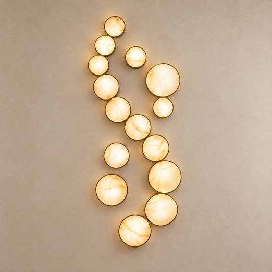 A modern wall-mounted light fixture composed of multiple oval and round stone alabaster glowing  orbs in warm cream and gold tones, arranged in a vertical spiral pattern against a textured beige wall.
