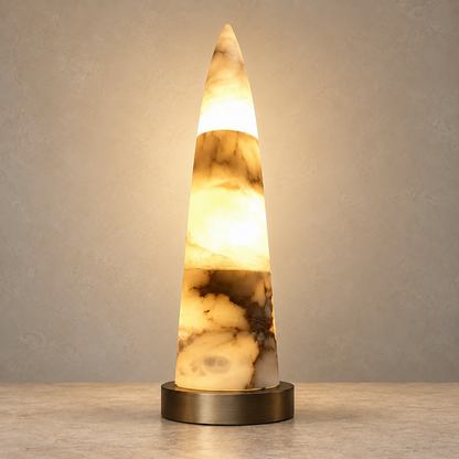 A glowing alabaster cone lamp with natural marbled patterns in cream, brown, and black, sitting on a round brushed brass base against a textured beige wall.