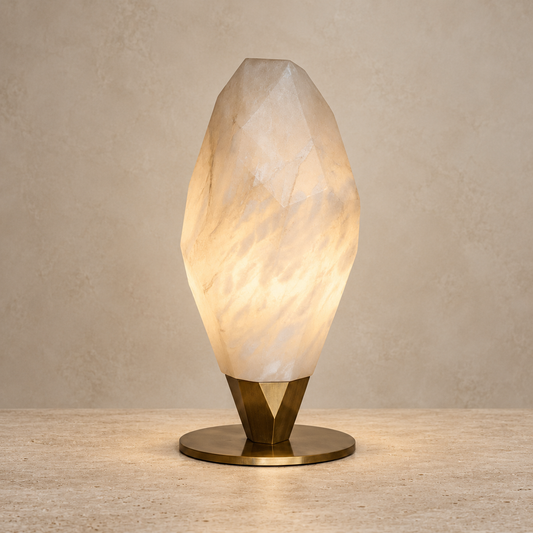 A faceted crystal-style table lamp with a glowing warm white and cream stone body on a polished brass base, set against a textured beige wall and stone surface.